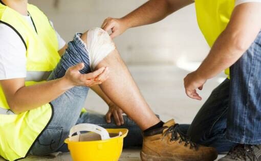 A worker helps another injured worker sitting on the ground with their knee bandaged